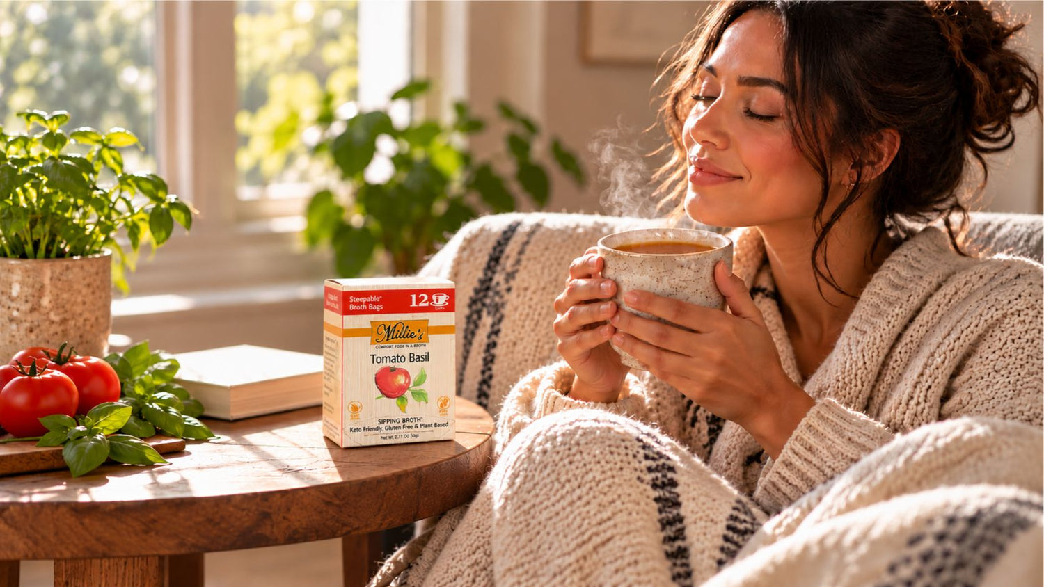 A woman in a cozy knit cardigan sits in a sunlit living room, holding a steaming mug. A box of Tomato Red herbal tea sits on the wooden table beside her, with fresh tomatoes and basil nearby.